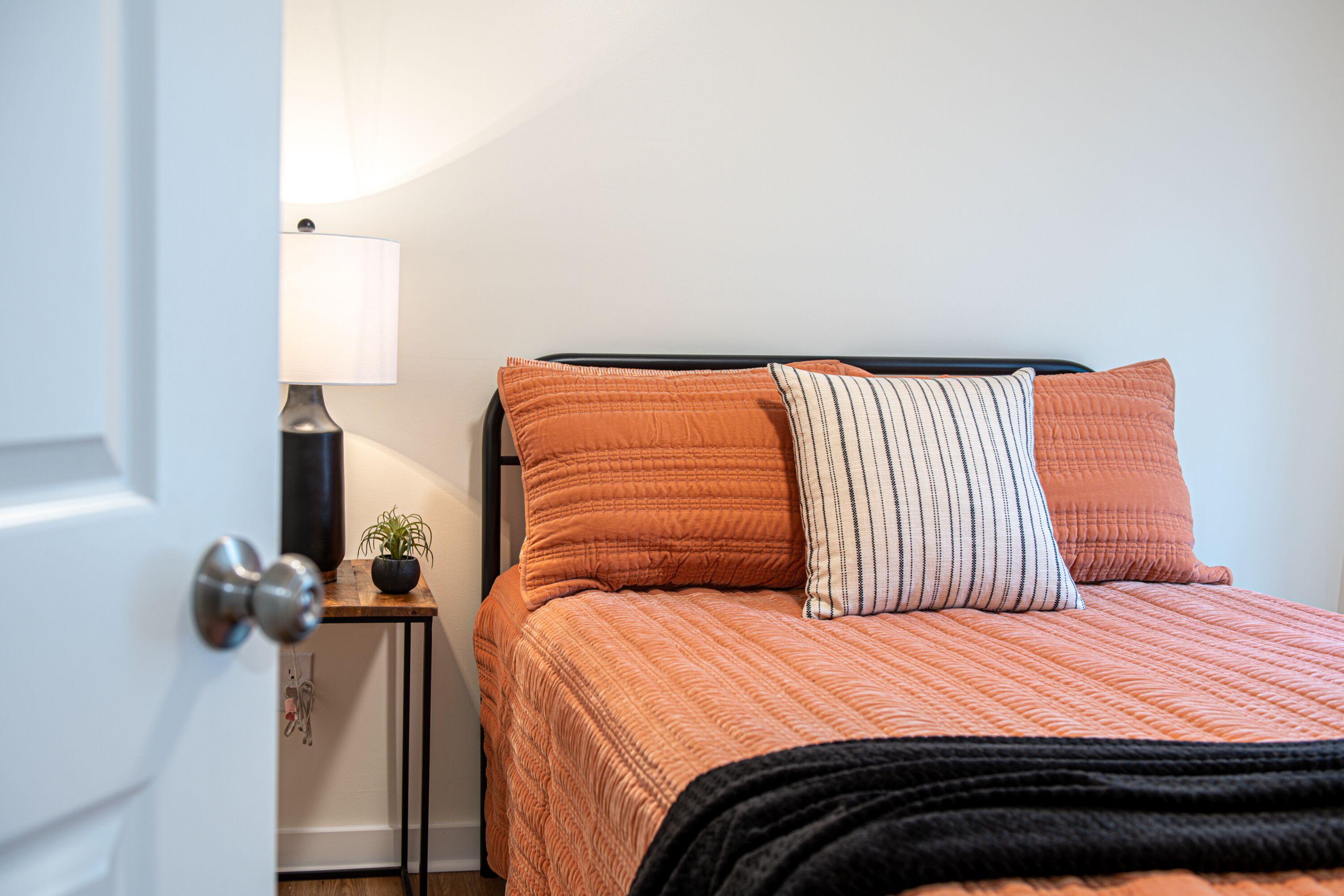 Una cama bien hecha con un edredón naranja, un cabecero negro y dos almohadas. Hay una mesita de noche con una lámpara y una planta en maceta.