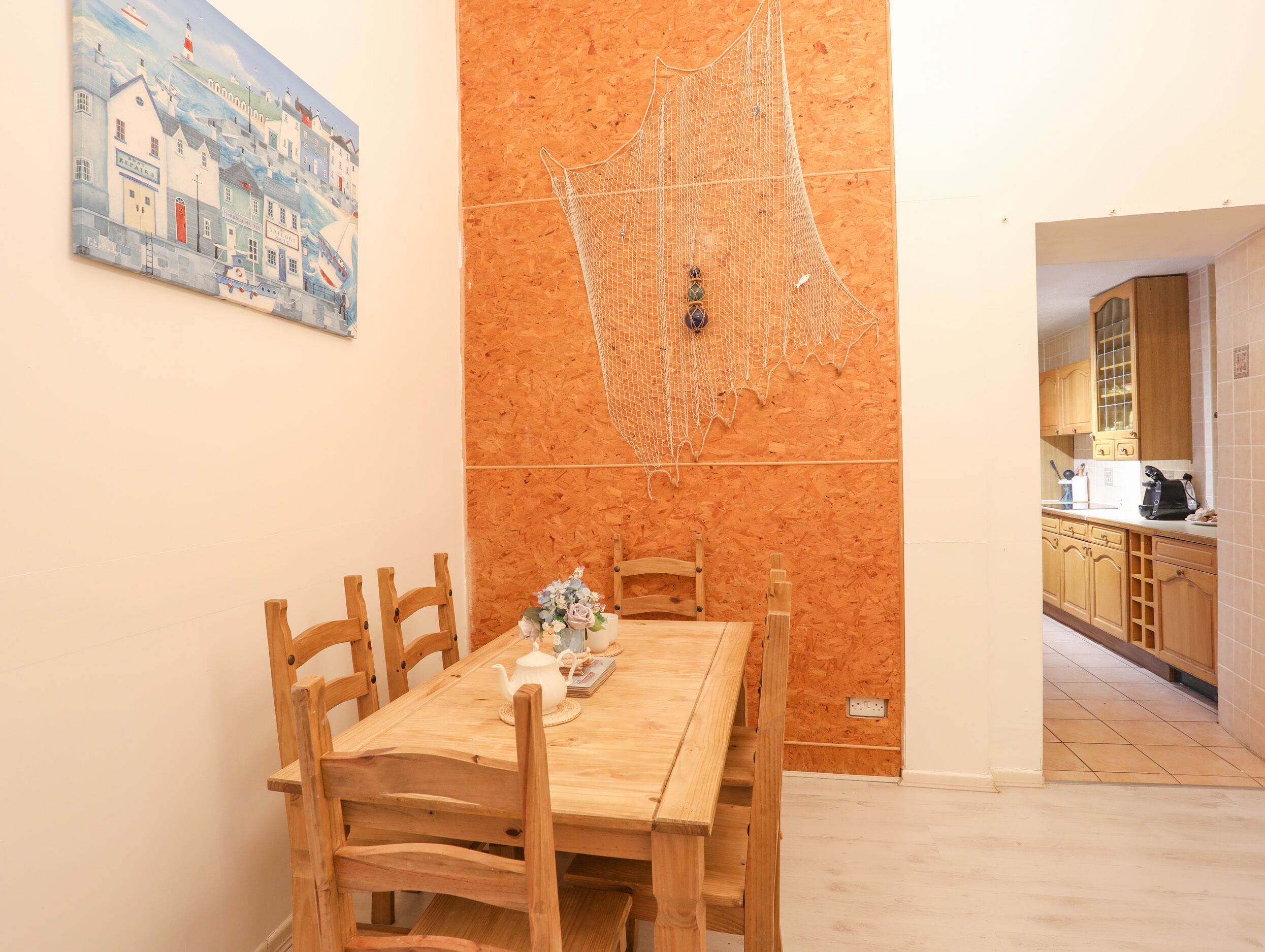 A dining area with a wooden table and chairs, a painting on the wall, and a kitchen visible through an open doorway.