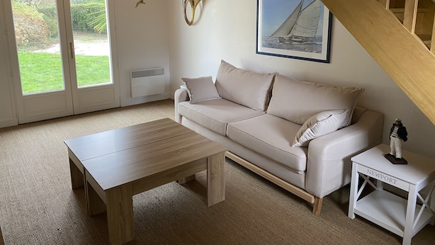 Ett vardagsrum med en beige soffa, ett soffbord i trä, ett vitt sidobord med en liten statyett, en inramad tavla med ett segelbåtsmotiv och en fågeldekoration.