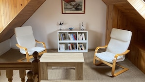 Ett mysigt rum på vinden med träribbor i taket, två vita fåtöljer, ett litet träbord och en bokhylla med böcker och dekorativa föremål.
