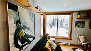 Un salon avec un grand tableau, un canapé, une fenêtre donnant sur des arbres couverts de neige et une petite table sur laquelle est posée une plante.