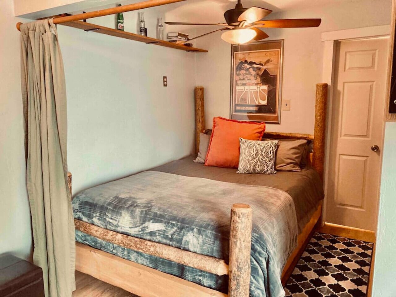 Een slaapkamer met een houten bedframe, een bed met een grijs- en blauw geborduurd sprei, een houten hoofdbord en een plafondventilator.