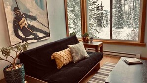 Un salon chaleureux avec un canapé noir, une petite table basse et une grande fenêtre donnant sur une forêt enneigée.