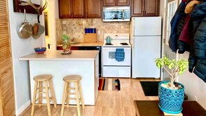 Une cuisine de taille réduite, dotée de meubles en bois, d’un four à micro-ondes, d’une cuisinière et d’un évier. On y trouve deux tabourets au comptoir et une plante en pot posée sur le sol.