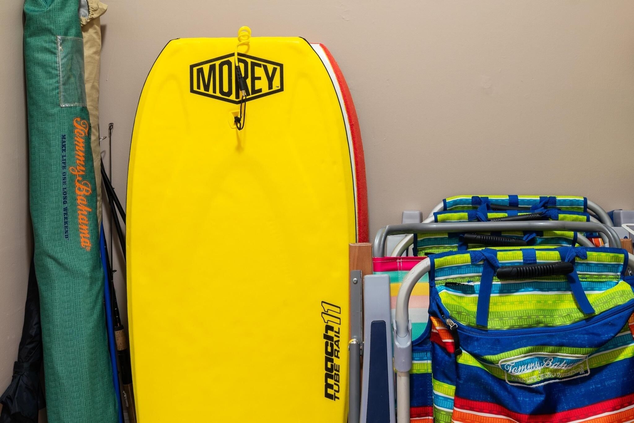 Ein gelbes Surfbrett mit 'MONEY'-Branding, eine grüne Tasche und eine blaugrüne Tasche mit dem Aufdruck 'Tommy's Bake'.