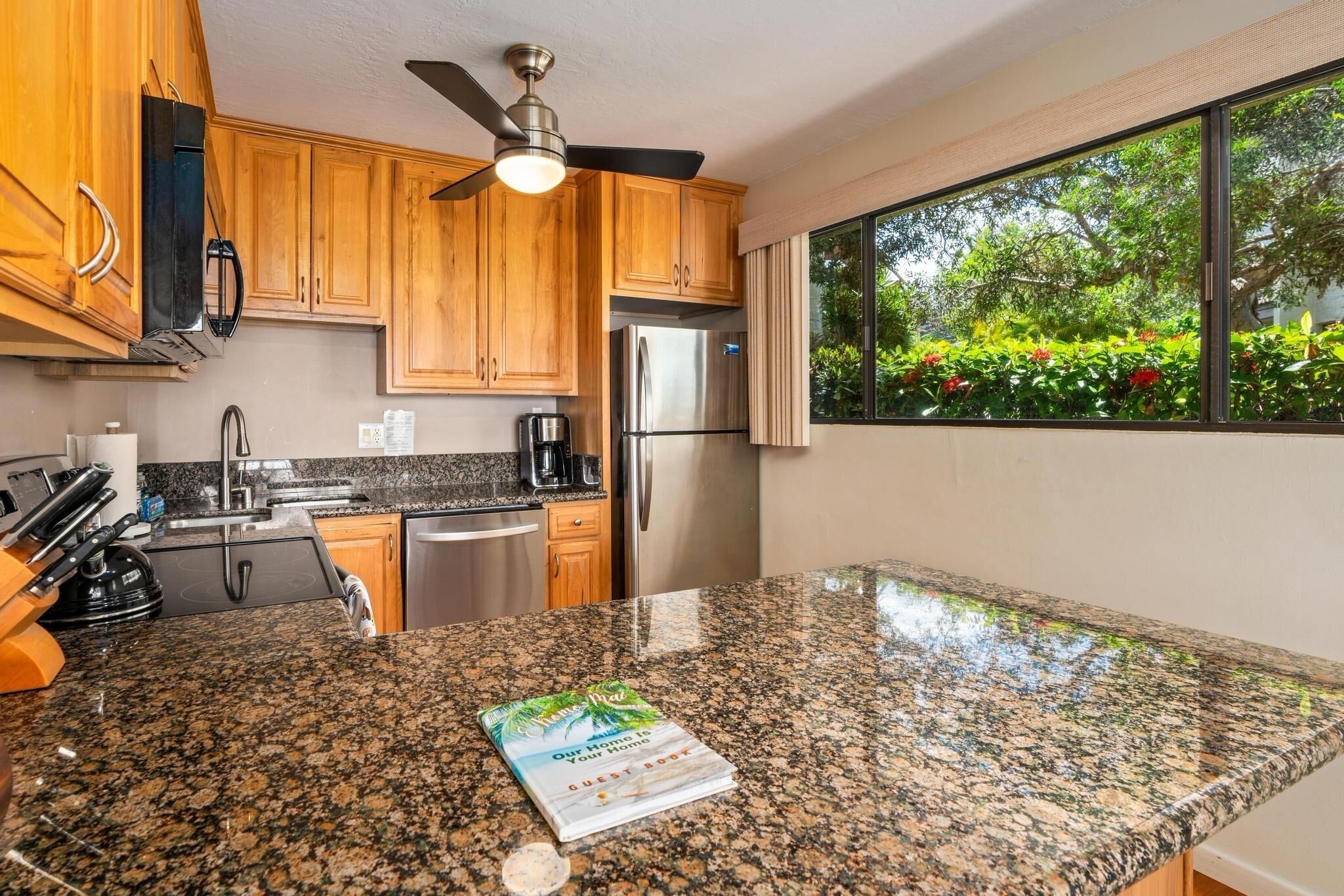 A modern kitchen with granite countertops, wooden cabinets, stainless steel appliances, and a large window offering a view of greenery.