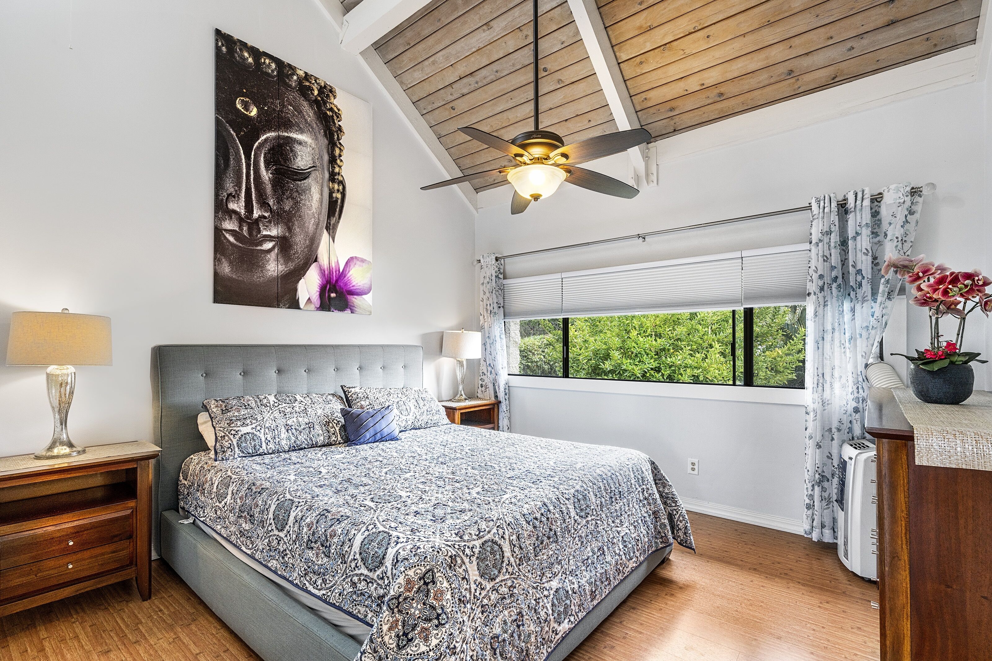 Un dormitorio con una cama grande, un ventilador de techo, una ventana con cortinas y un cuadro de Buda en la pared.