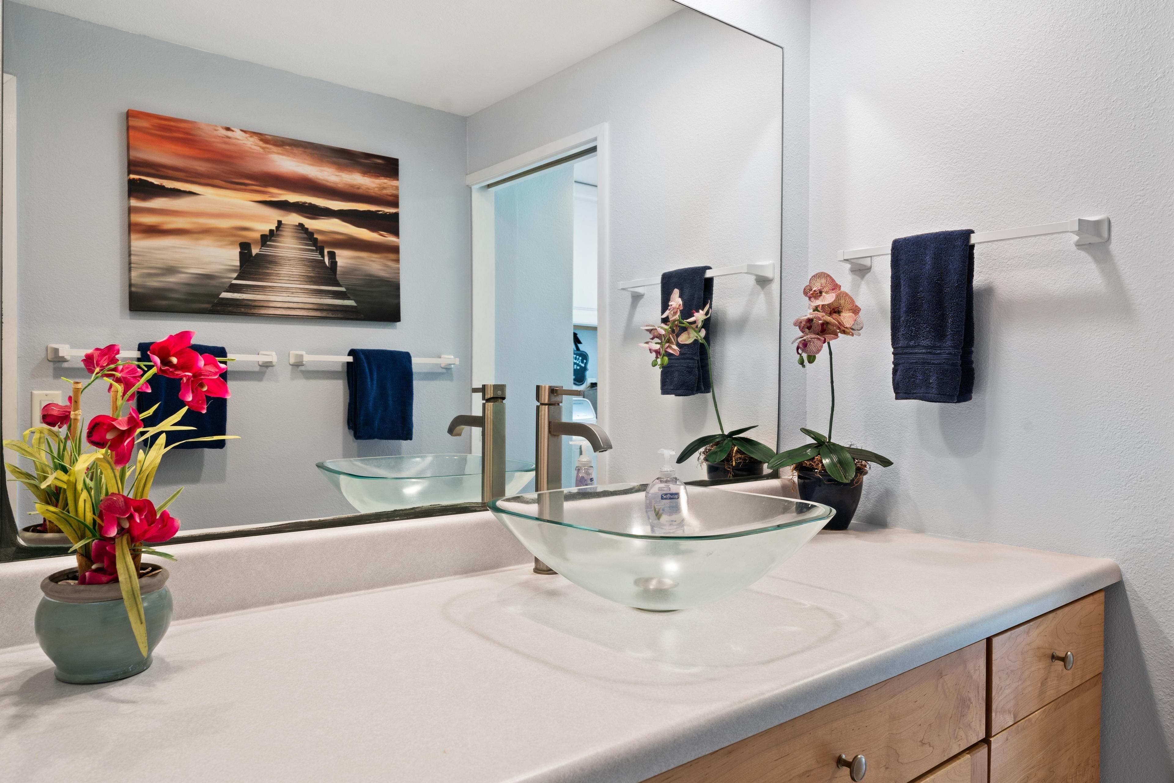 Un baño moderno con un lavabo de vidrio, una planta en maceta y un cuadro en la pared.