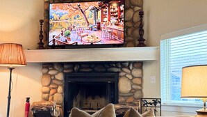 Een gezellige woonkamer met een stenen open haard, een televisie erboven en een comfortabele zithoek met een bank en kussens.