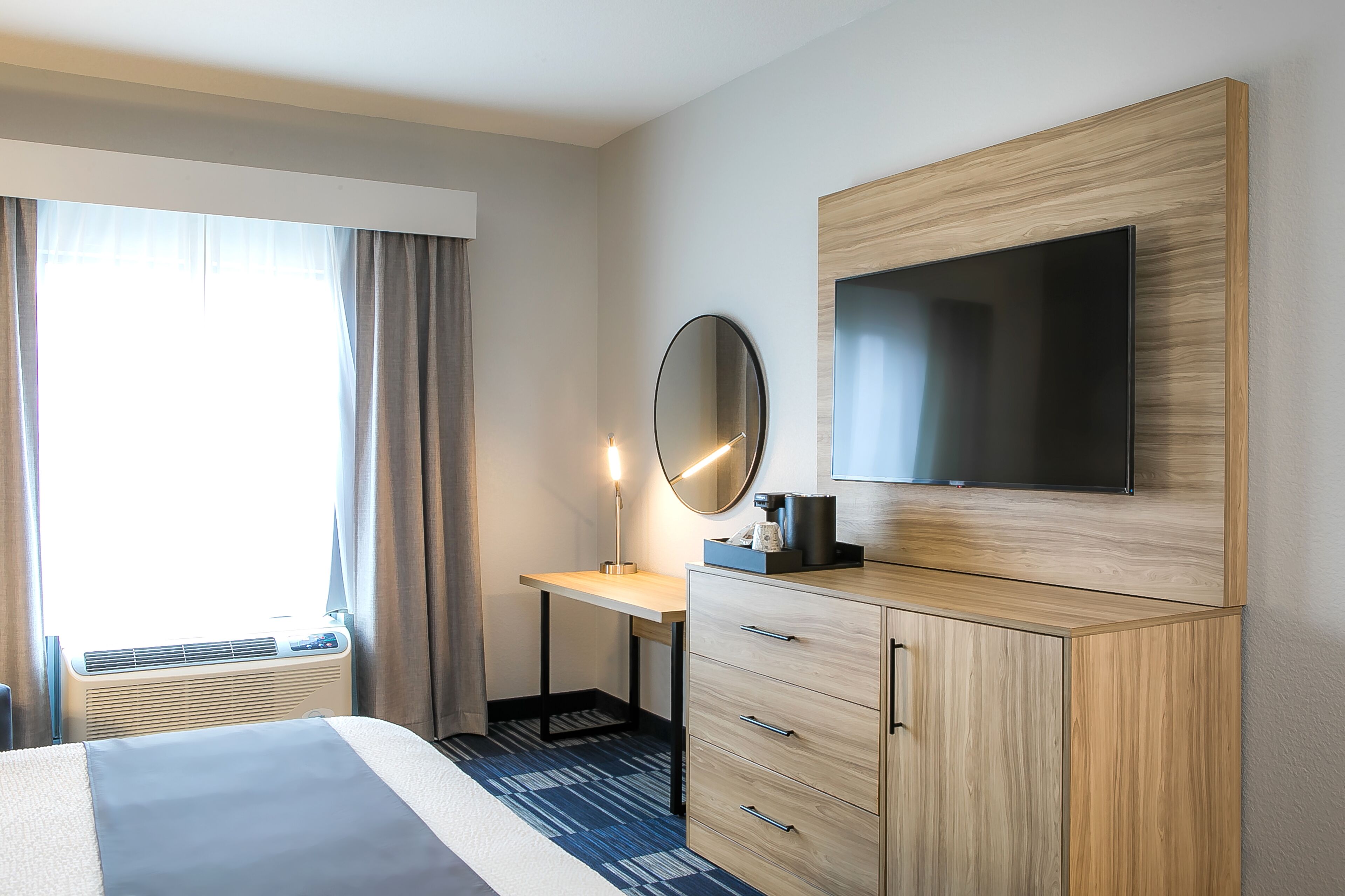 Una habitación de hotel con cama, escritorio con silla, televisor, espejo y armario.