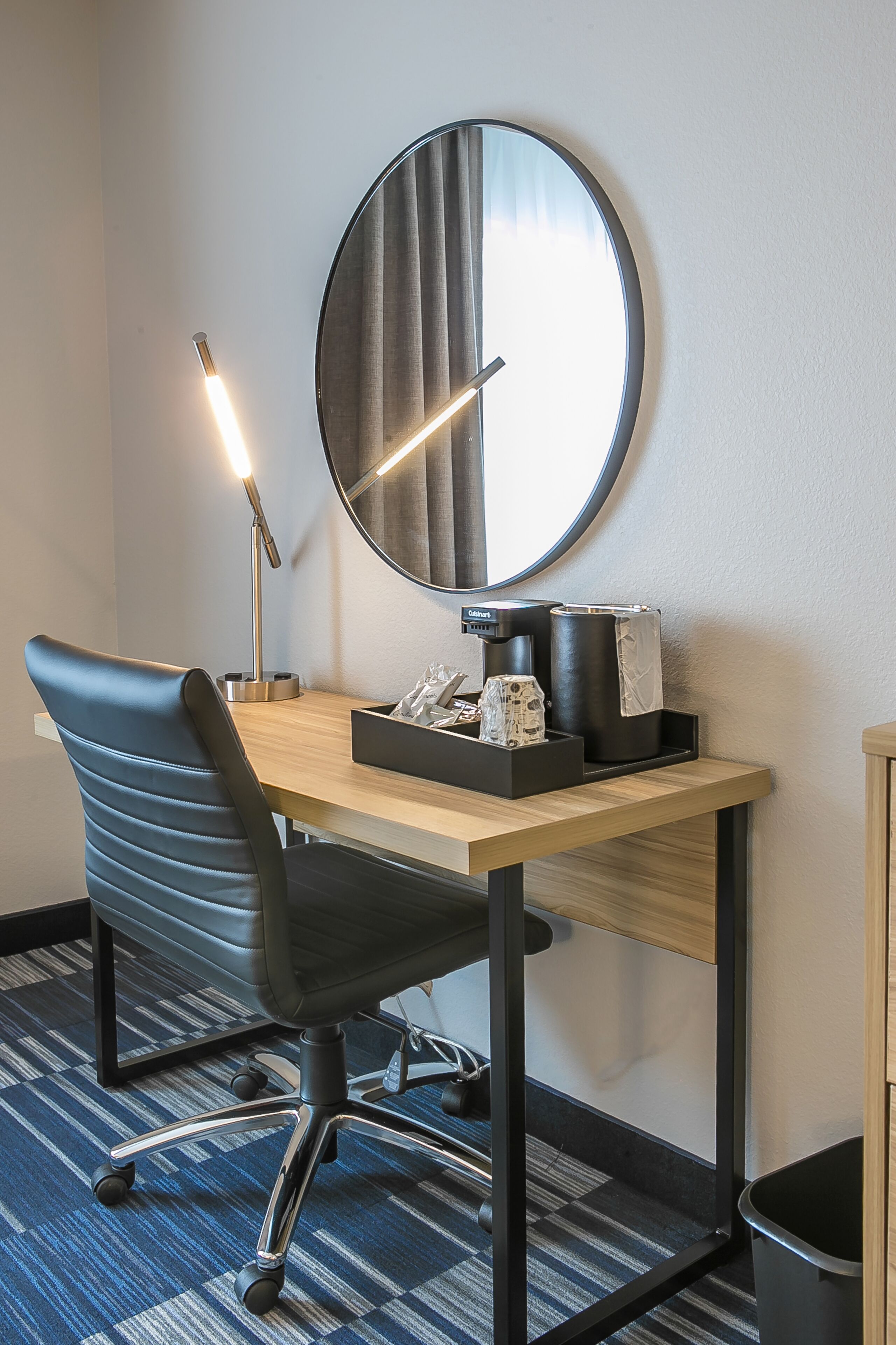 A hotel room with a desk, a chair, a round mirror, a coffee maker, and a trash bin.