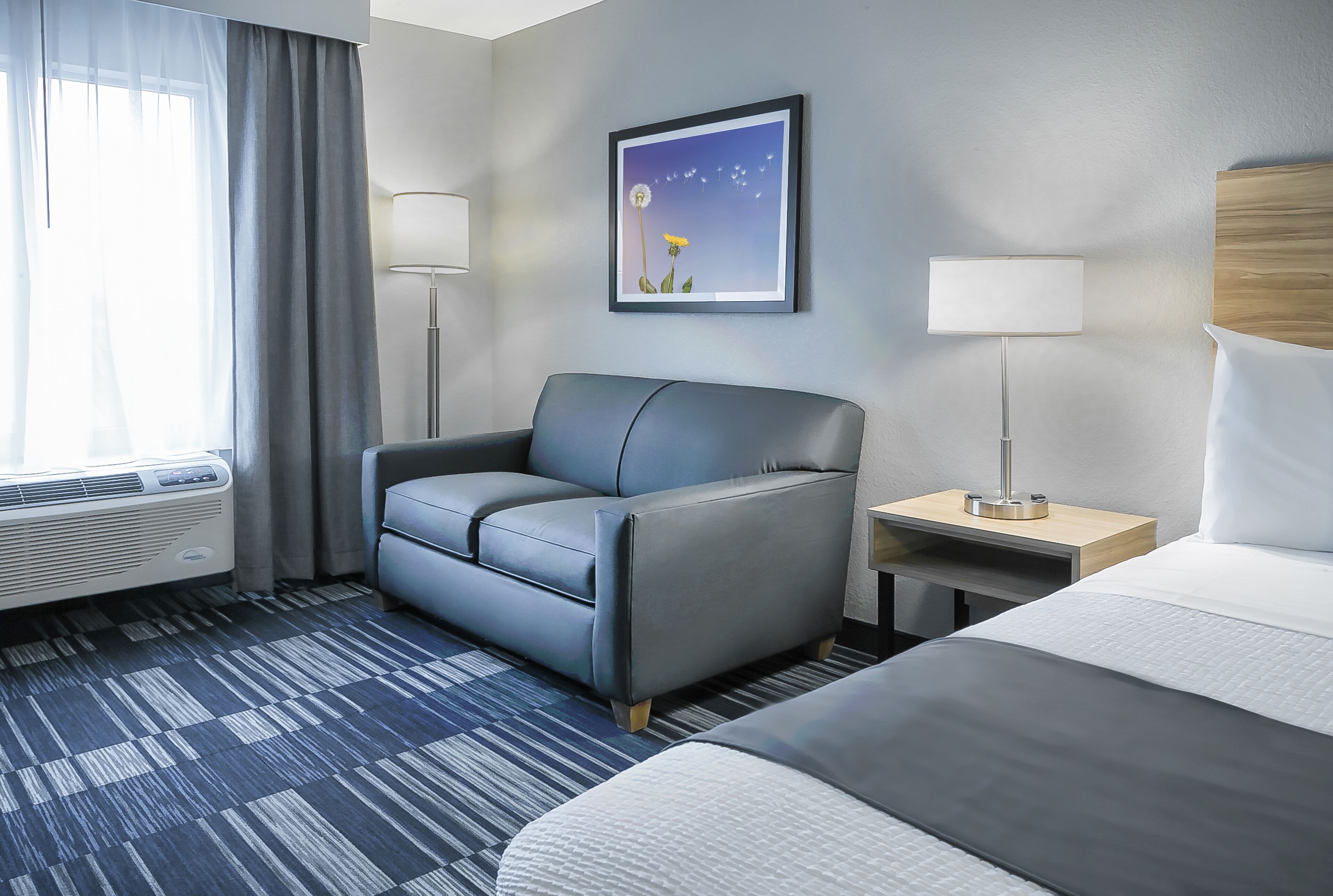 Una habitación de hotel con una cama, un sofá gris, una mesita de noche con una lámpara, un cuadro en la pared y una ventana con cortinas.