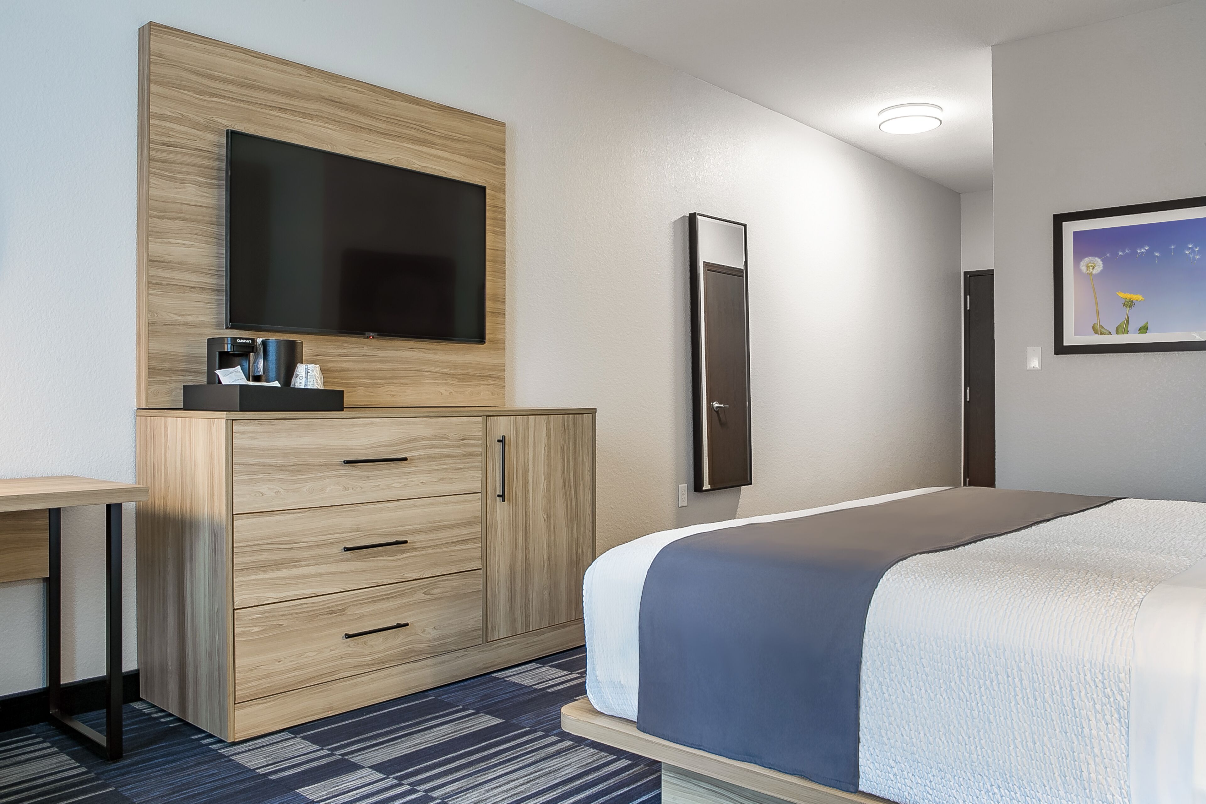 A modern hotel room with a large bed, a flat-screen TV mounted on a wooden cabinet, a desk with a chair, and a framed picture on the wall.
