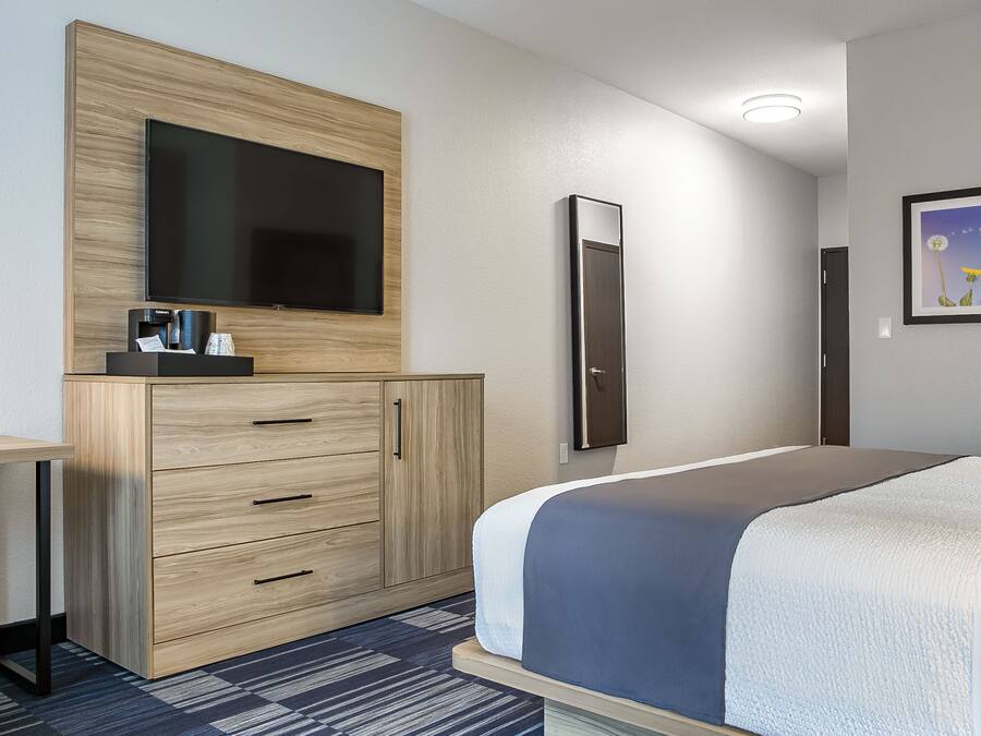 A modern hotel room with a large bed, a flat-screen TV mounted on a wooden cabinet, a desk with a chair, and a framed picture on the wall.