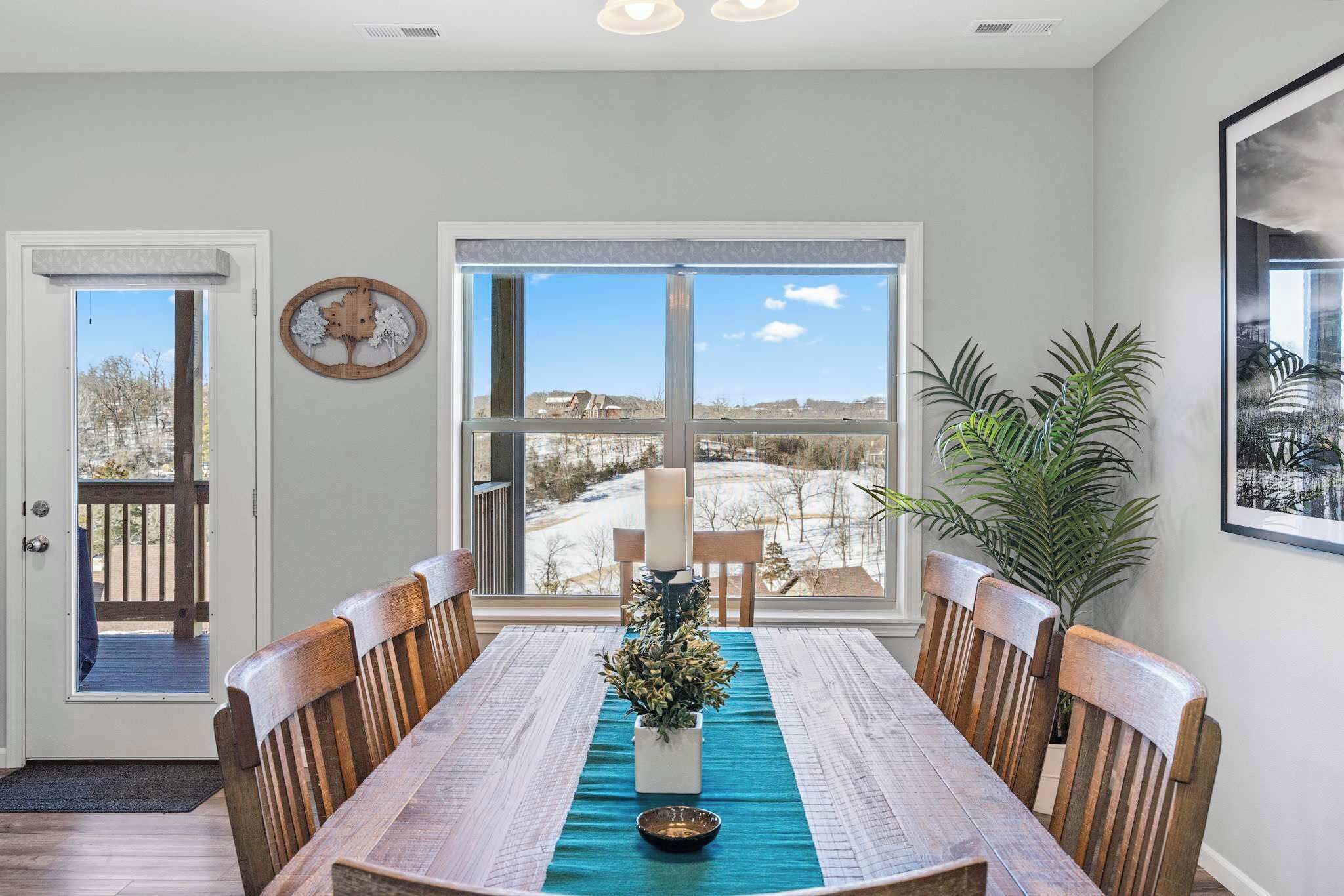 A dining area with a wooden table, chairs, a vase with flowers, and a view of a snowy landscape through large windows.