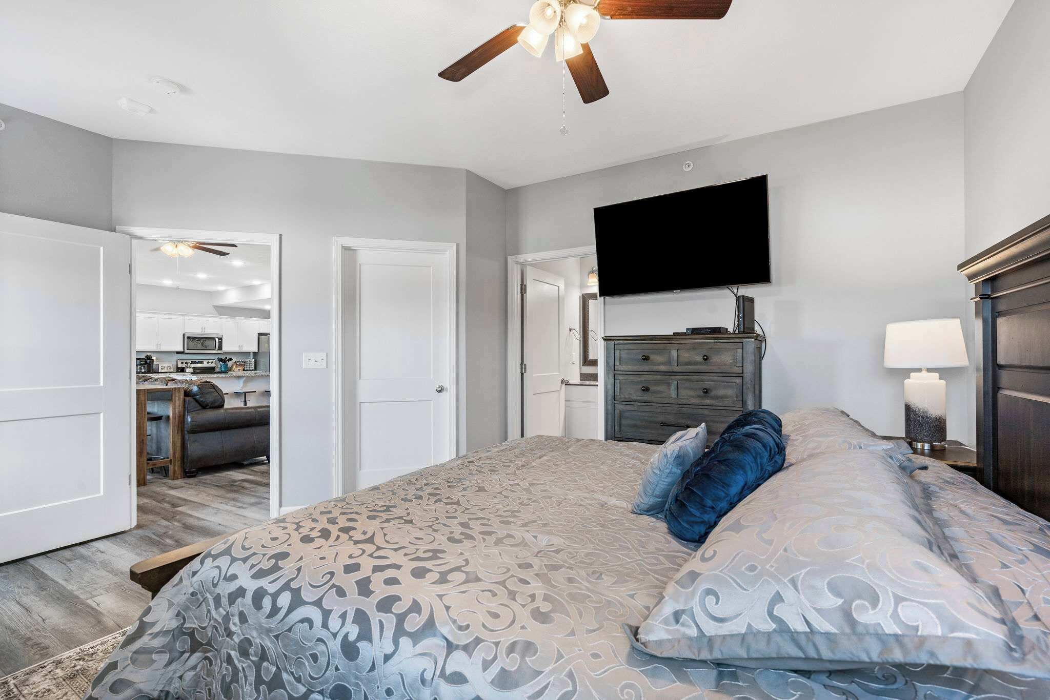 A bedroom with a patterned bedspread, a ceiling fan, a TV mounted on the wall, and a dresser with a lamp.