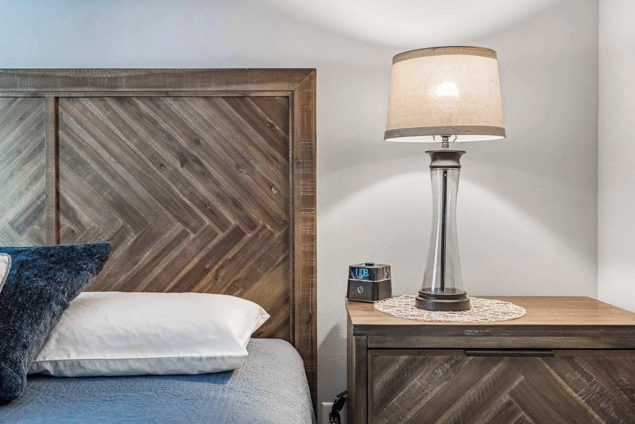 Una cama bien hecha con tablero de madera, una lámpara de noche y un reloj pequeño sobre una mesita de noche de madera.