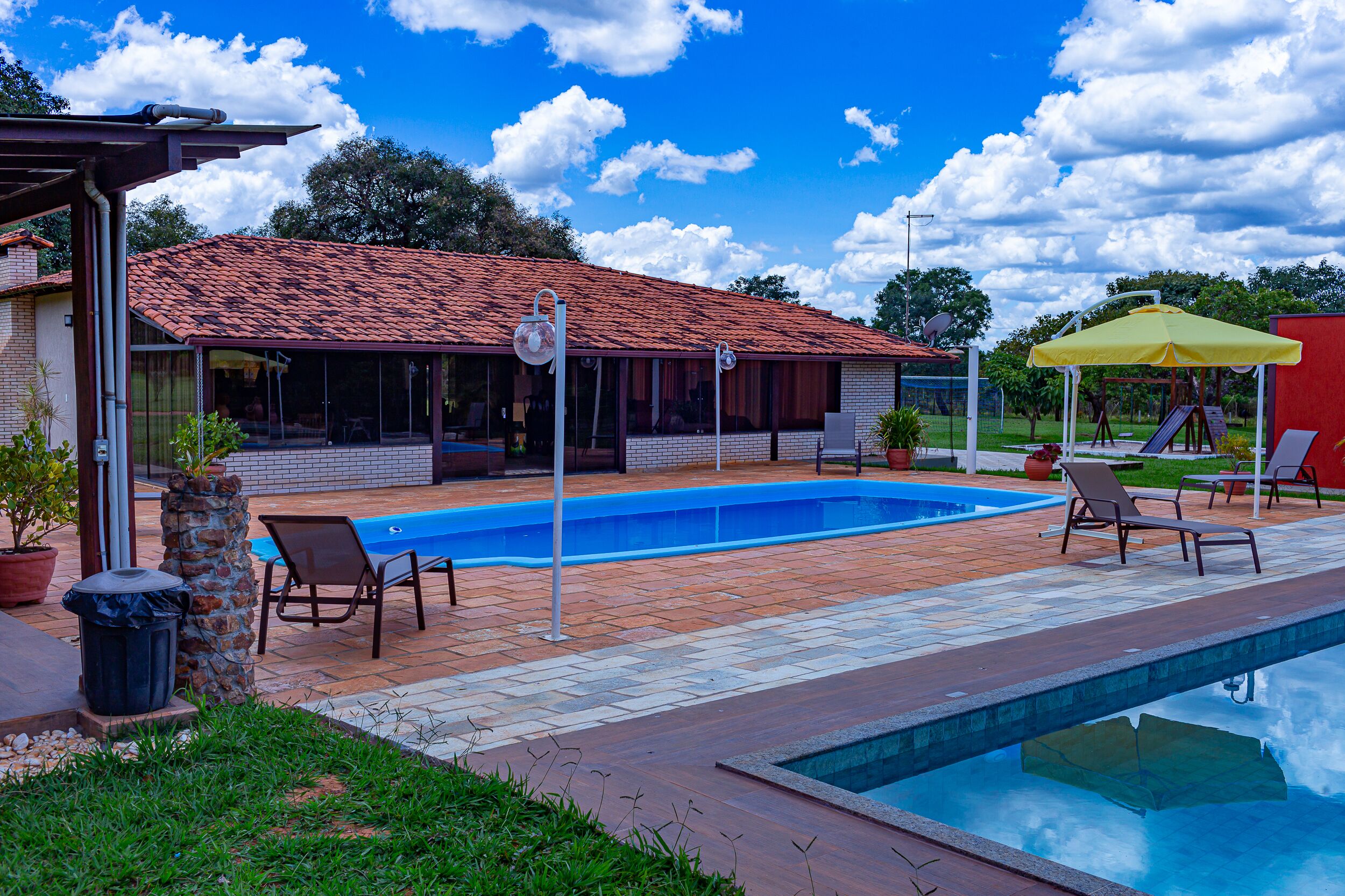 Una piscina con un deck de azulejos, sillas de descanso y un patio cubierto, con un edificio de techo rojo al fondo.