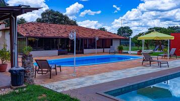 Una piscina con un deck de azulejos, sillas de descanso y un patio cubierto, con un edificio de techo rojo al fondo.