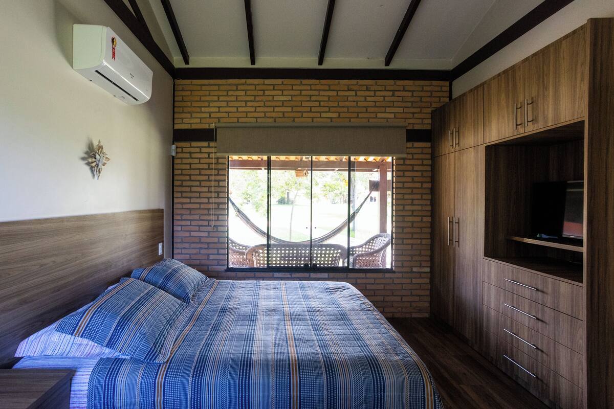 A bedroom with a bed, a TV cabinet, and a hammock by a brick wall window.