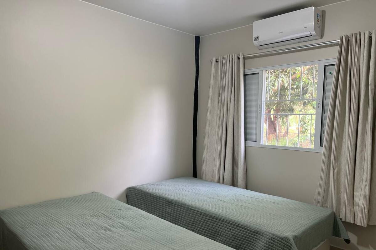 A room with two single beds, a window with curtains, and an air conditioning unit.