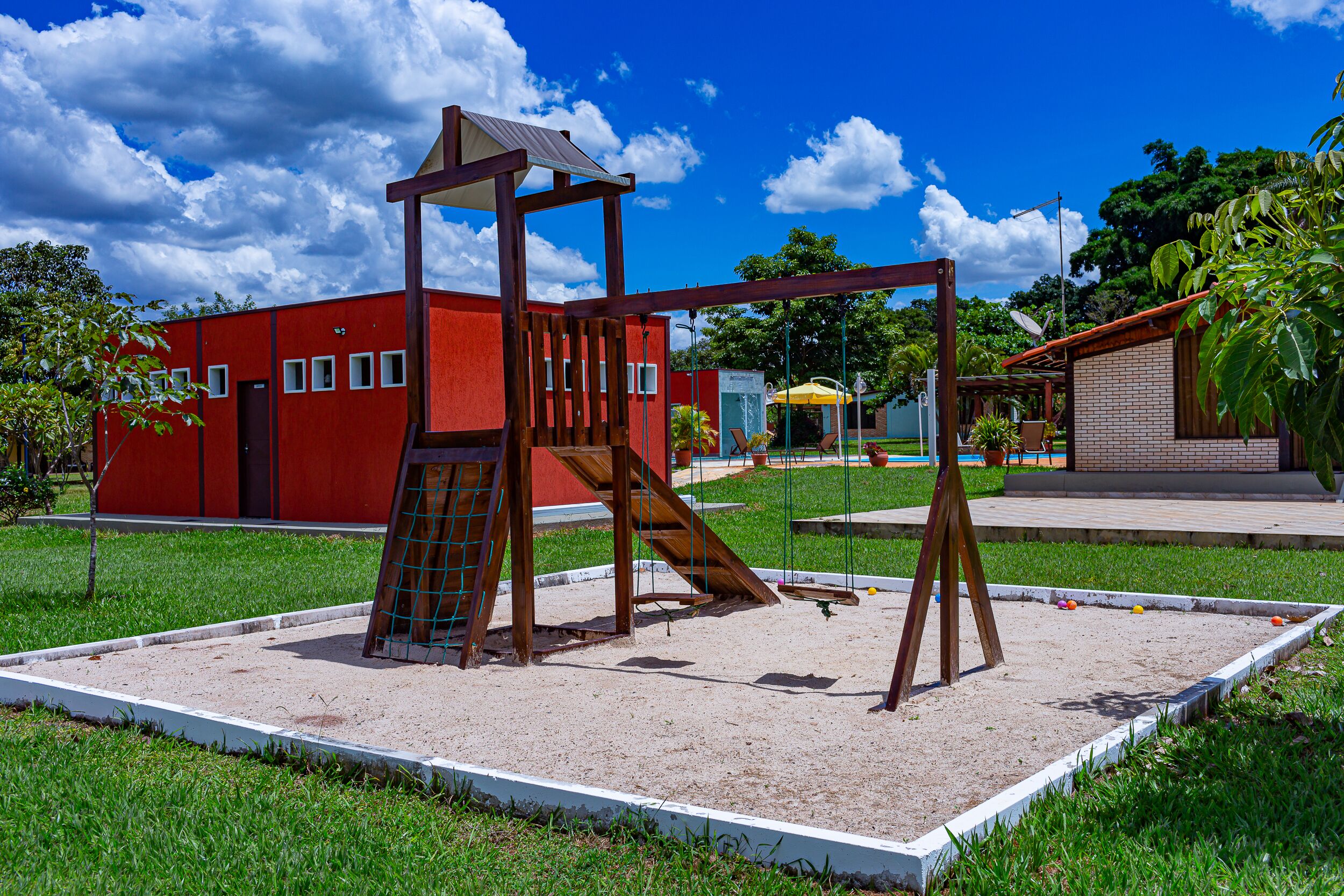Un parque infantil con columpios y una estructura de escalada, rodeado por una valla, con un edificio rojo al fondo.