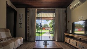 A living room with a sofa, a coffee table, and a TV on a wooden stand. There is a sliding glass door leading to a patio with a lawn and a red car in the background. - Villaggio dos Pássaros (Paranoa)