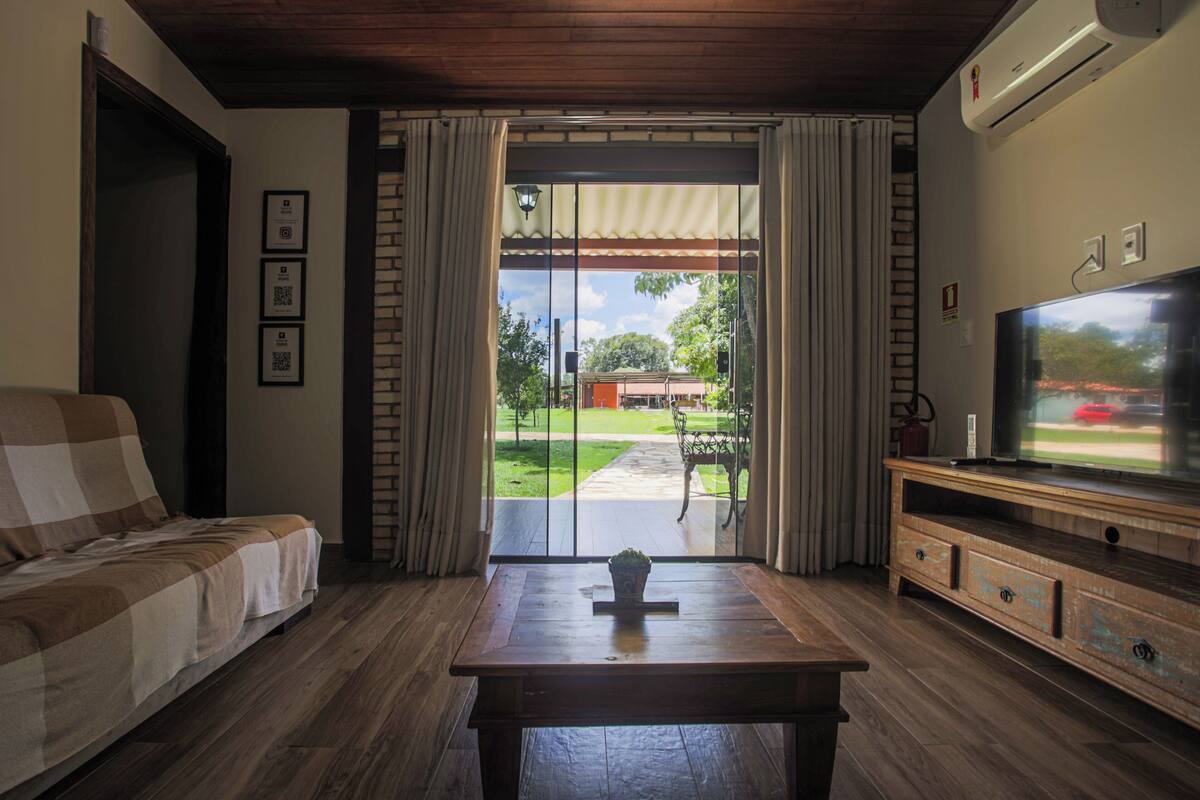 A living room with a sofa, a coffee table, and a TV on a wooden stand. There is a sliding glass door leading to a patio with a lawn and a red car in the background.