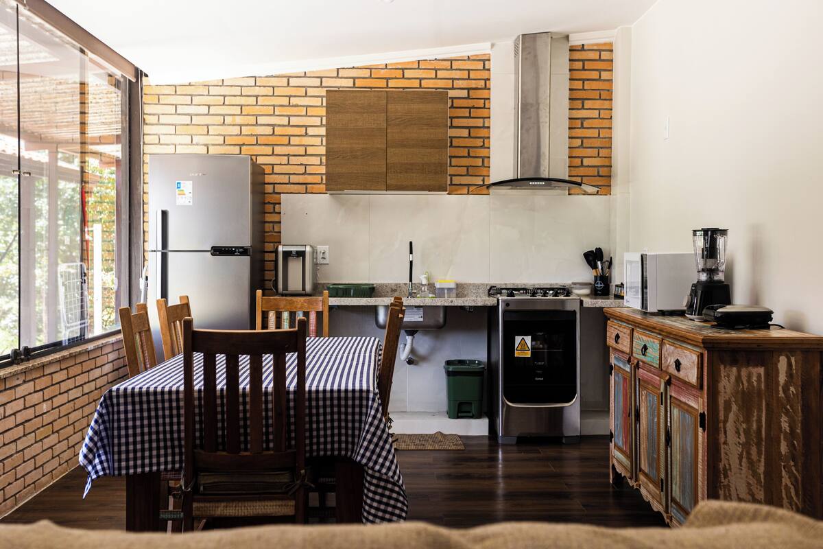 A compact kitchen with a refrigerator, stove, and sink, a dining table with chairs, and a brick wall.