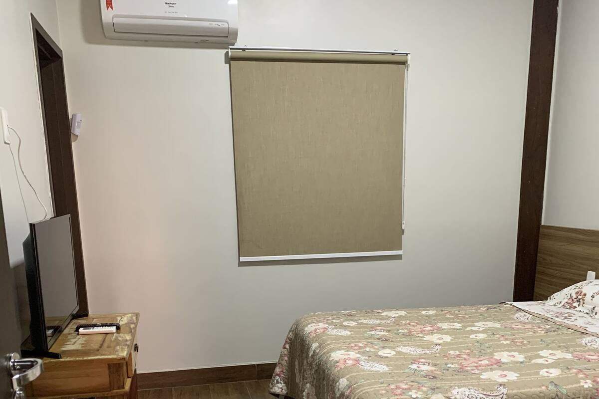 A bedroom with a bed, a desk, a chair, a window with a blind, and an air conditioning unit on the wall.