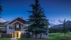 A two-story house with a prominent evergreen tree in front, a well-maintained lawn, and a mountainous backdrop.
