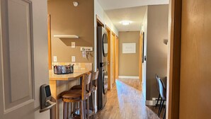 A narrow hallway with a kitchen area on the left, featuring a wooden counter, bar stools, and a refrigerator.