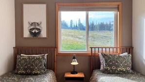 A bedroom with two beds, a window with a view of a forest, and a raccoon picture on the wall.