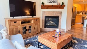 A living room with a flat-screen TV, wooden entertainment unit, fireplace, and a kitchen visible in the background.