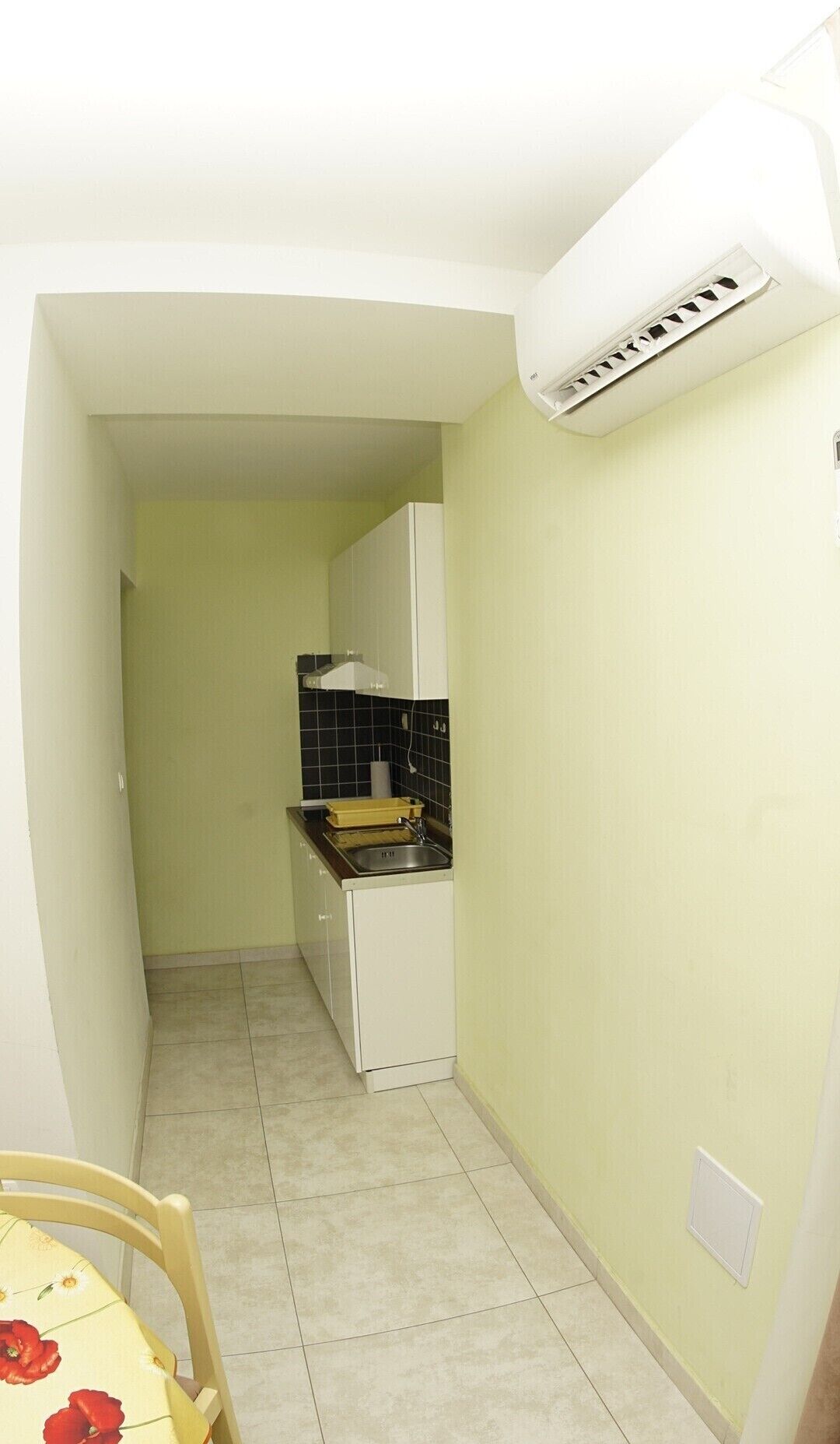 A narrow kitchen with white cabinets, a black tiled backsplash, and a white air conditioning unit mounted on the wall.