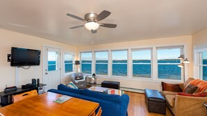 Ein Wohnzimmer mit großem Fenster und Meerblick, Deckenventilator, Wand-Fernseher und Esstisch mit Stühlen.