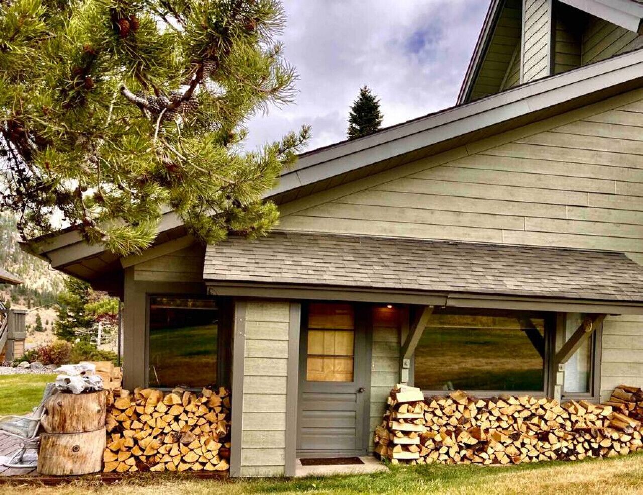 A modern house with a covered porch, a stack of firewood, and a pine tree.