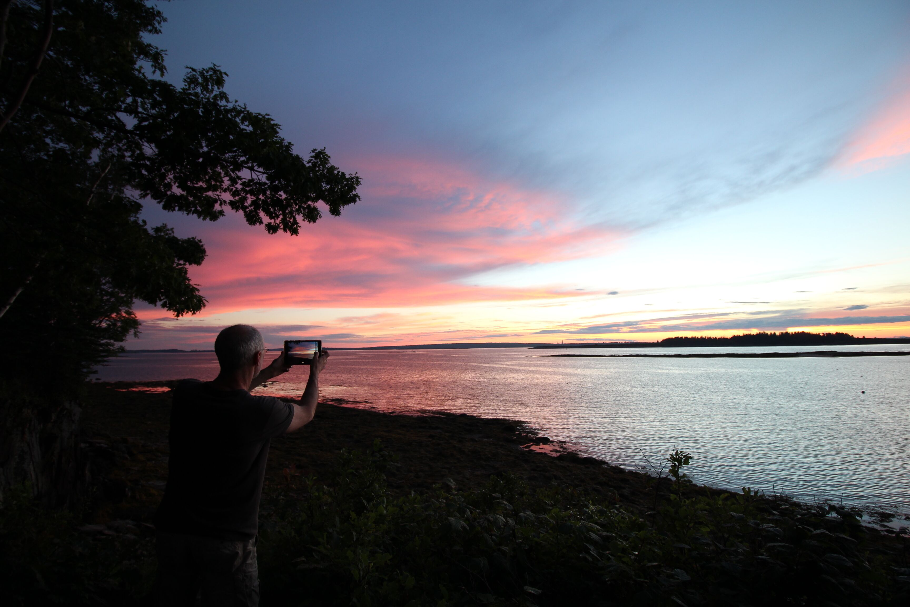 A person taking a photo of a sunset over a body of water with trees on the left.
