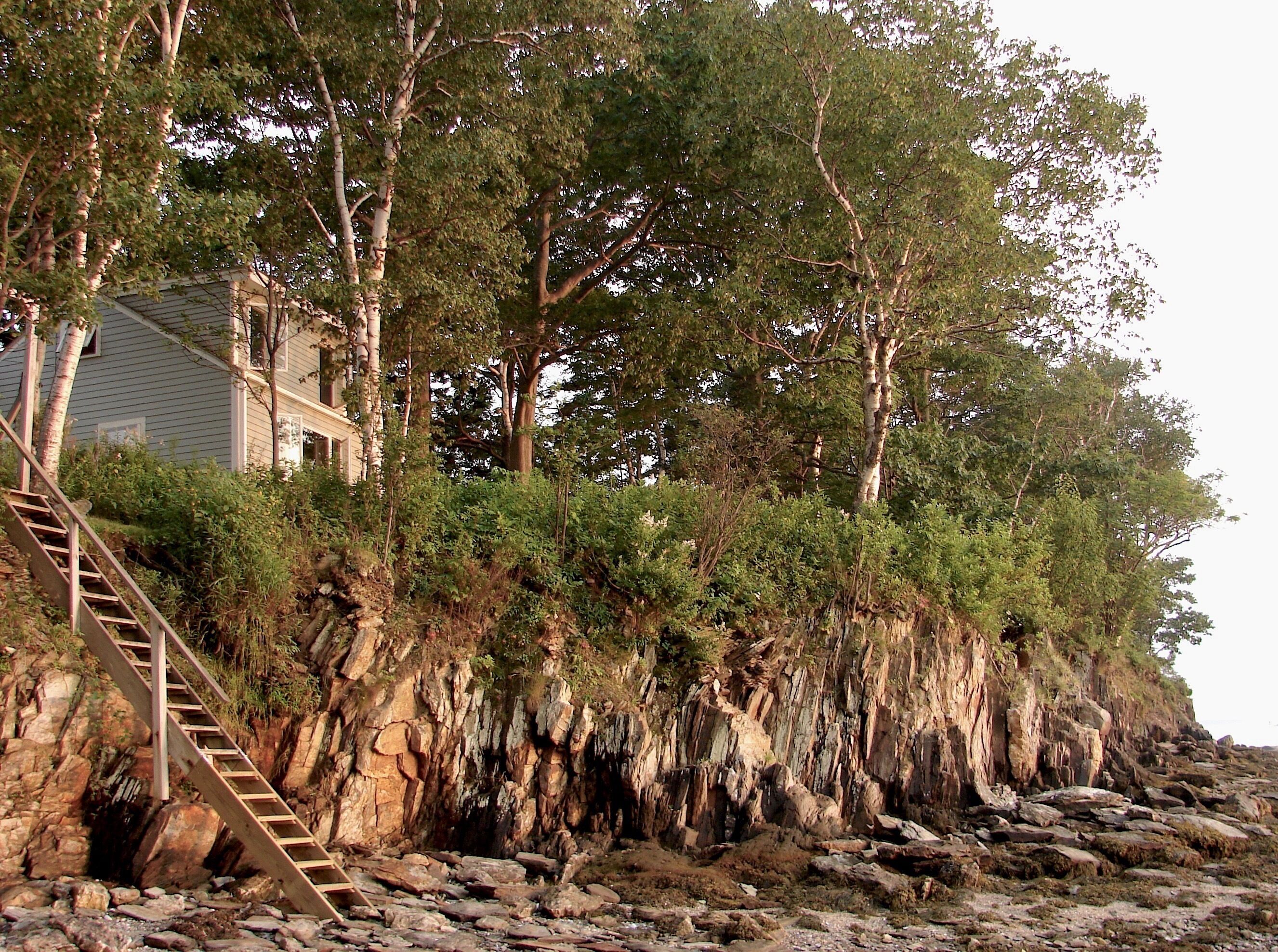 A coastal house with a wooden staircase leading up to a rocky cliff.