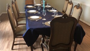 Een eetkamer met een rechthoekige tafel gedekt voor zes personen, omringd door zes stoelen. Een kroonluchter hangt aan het plafond, en een houten kast met glazen deuren bevindt zich op de achtergrond.