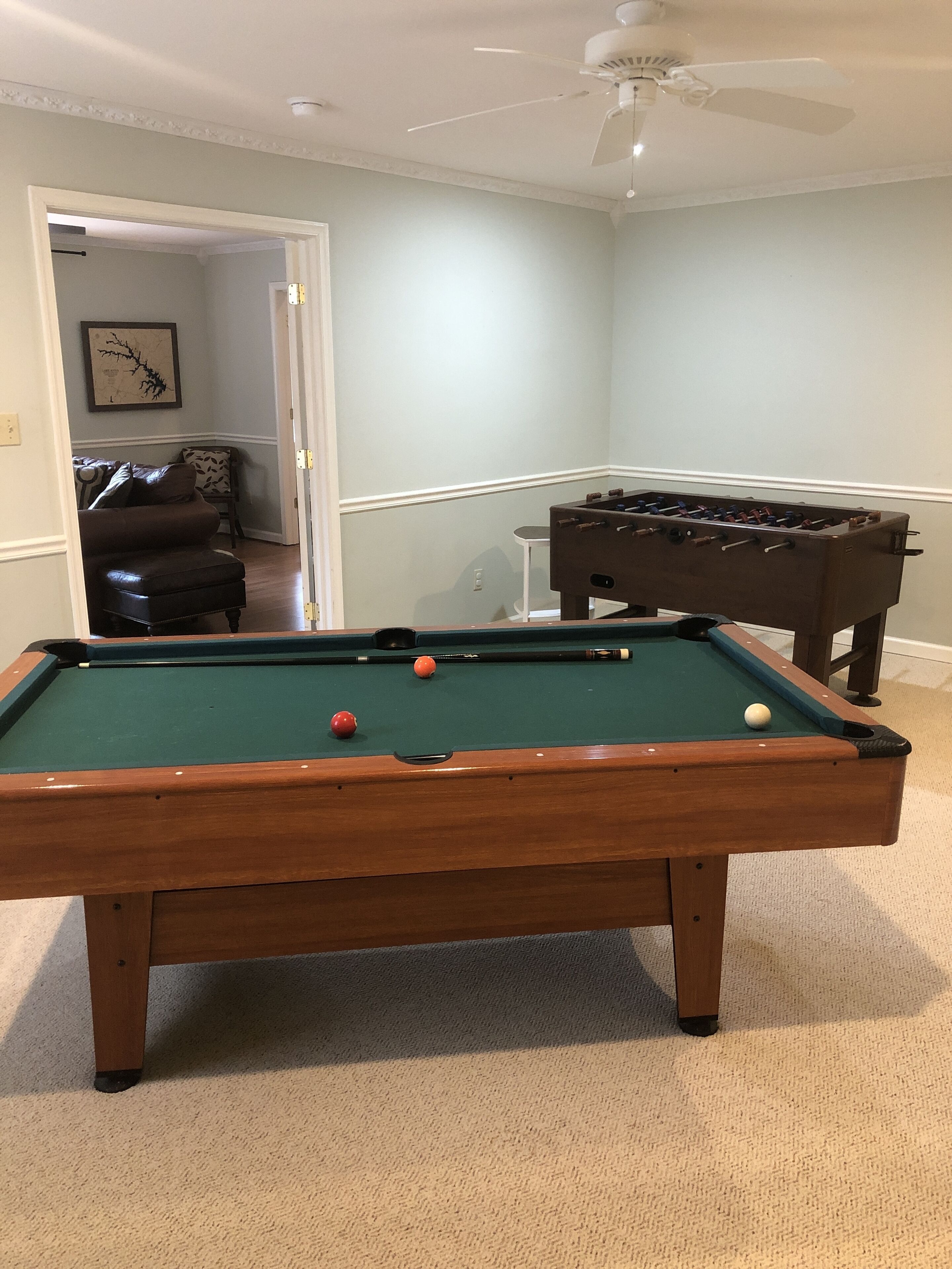 A pool table with balls, a foosball table, and a ceiling fan in a room with light-colored walls and carpet.