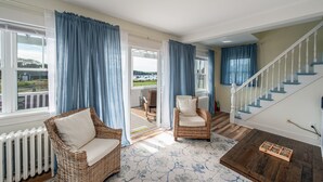 Ein geräumiges Wohnzimmer mit einem Rattan-Sofa, einem Sessel und Blick ins Freie durch große Fenster.
