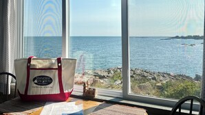 Vue depuis une pièce dotée d’une grande fenêtre donnant sur la mer, une plage et une côte rocheuse. Un sac orné d’un logo et d’une inscription est posé sur le rebord de la fenêtre.
