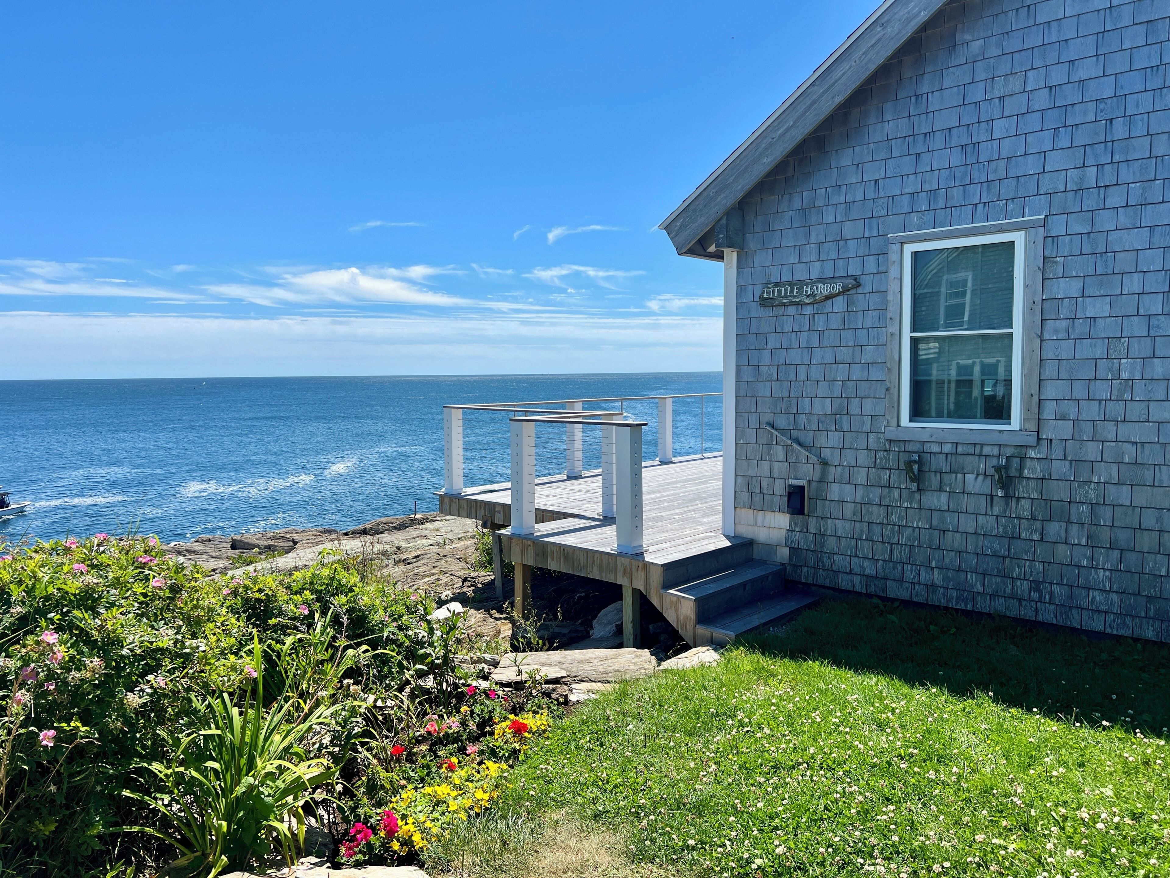 Ein Küstenhaus mit einer Holzdiele, einem kleinen Garten mit Blumen und Blick aufs Meer.