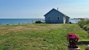 Une petite maison de plain-pied, dotée d’un toit à pignon et d’une cheminée, située sur un terrain herbeux et offrant une vue sur la mer.