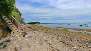Une plage rocheuse avec un escalier en bois descendant jusqu’à la rive, des bateaux sur l’eau et une côte lointaine bordée d’arbres.