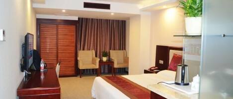 A hotel room with a bed, desk, chairs, a TV, a plant, and a window.