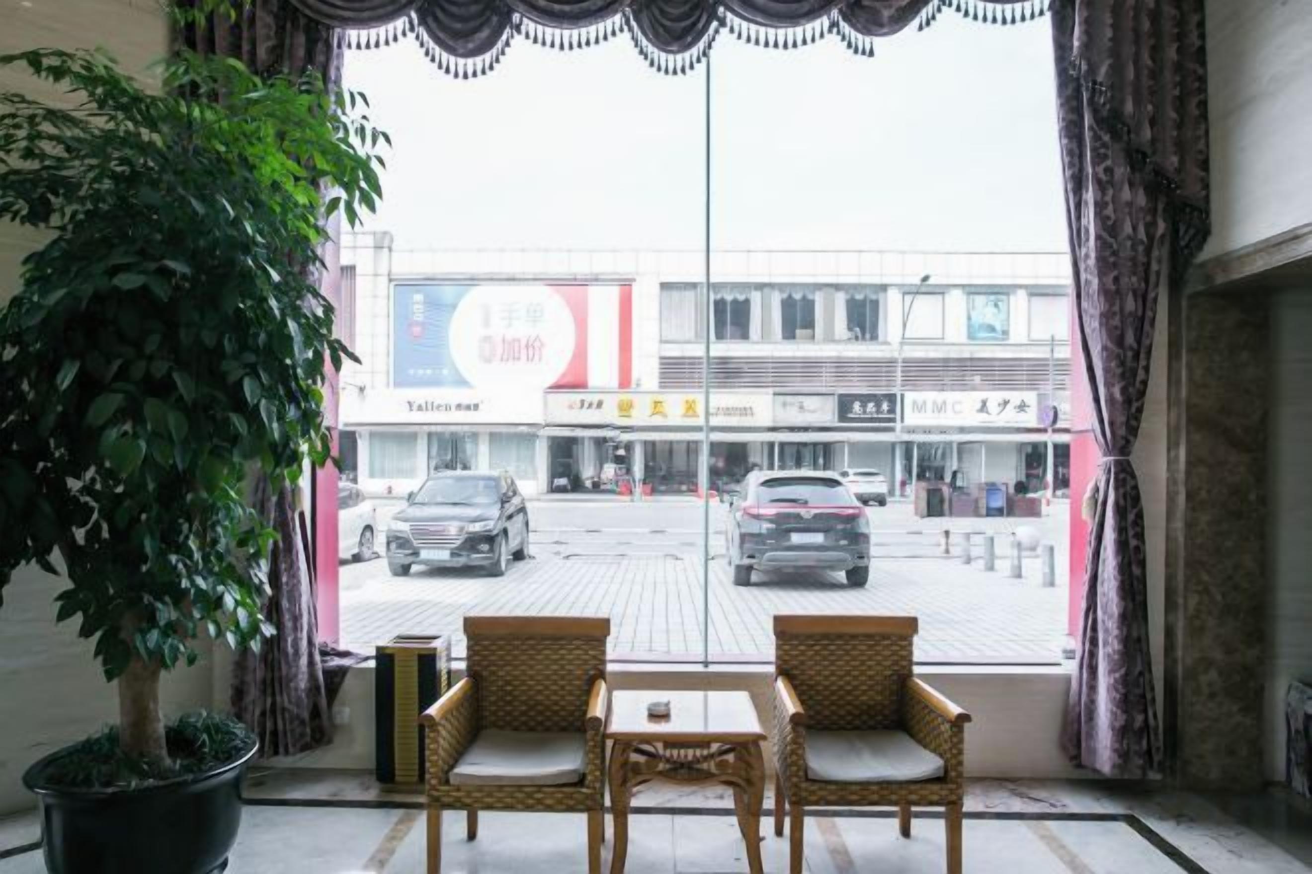A hotel room with a view of a street, two wicker chairs, a small table, and a potted plant.