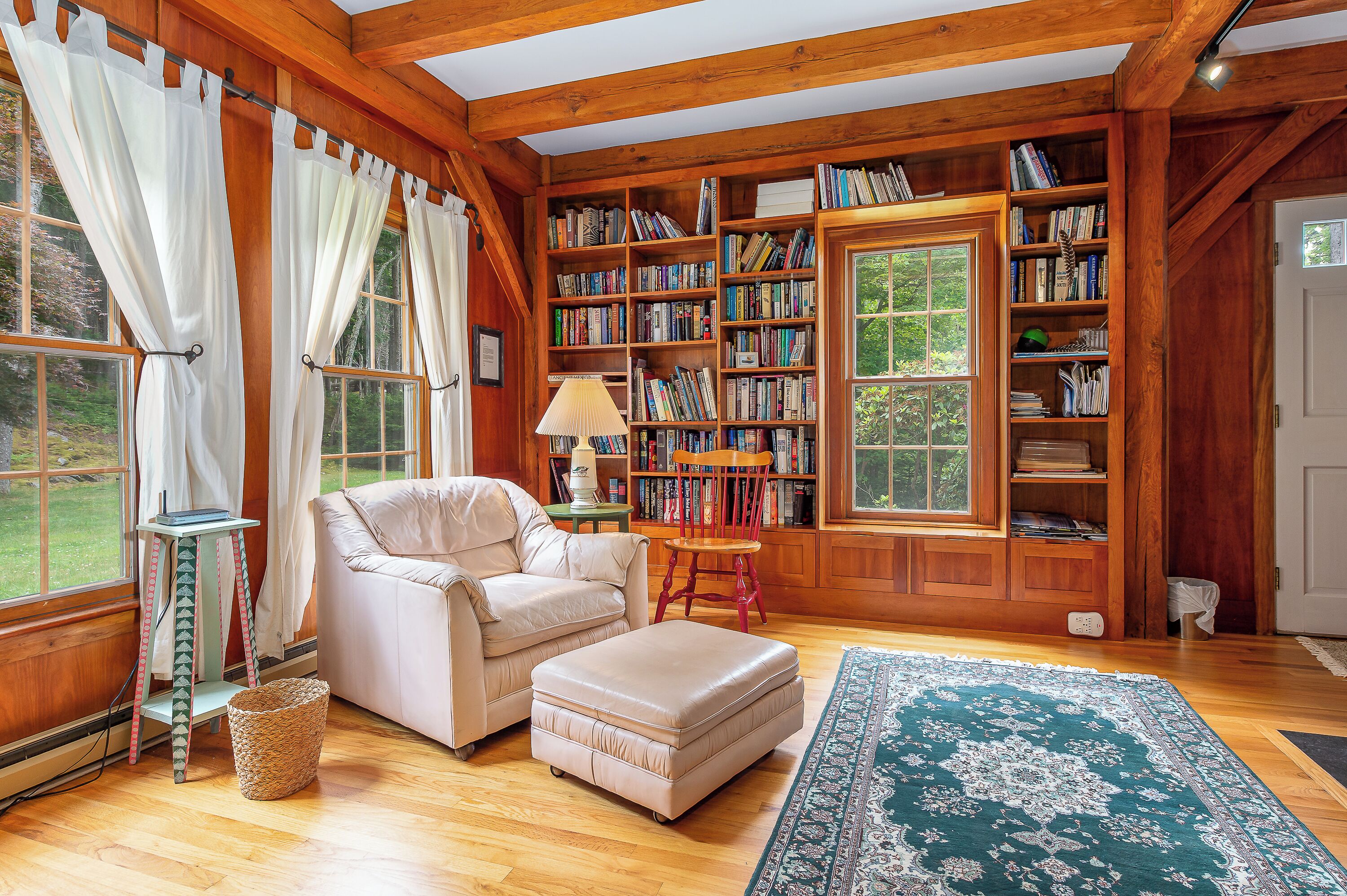 A cozy room with a wooden bookshelf, a beige armchair, a small table, and a large window with curtains.