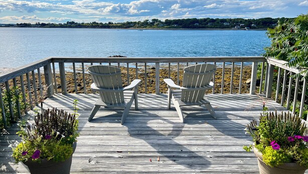 Une terrasse en bois avec deux fauteuils Adirondack donnant sur un lac paisible, le tout entouré de verdure et sous un ciel dégagé.